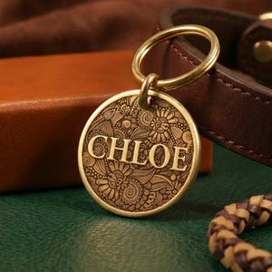May include: A circular brass pet tag featuring an intricate engraved floral and swirl pattern with the name CHLOE centered in bold capital letters. The tag is attached to a gold-toned metal split ring, showcasing a detailed metallic finish.