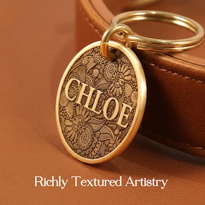 May include: A round gold-toned metal pet tag featuring an intricate engraved floral pattern and the name CHLOE in bold capital letters. The tag is attached to a gold-toned split ring and rests against a smooth brown leather surface.