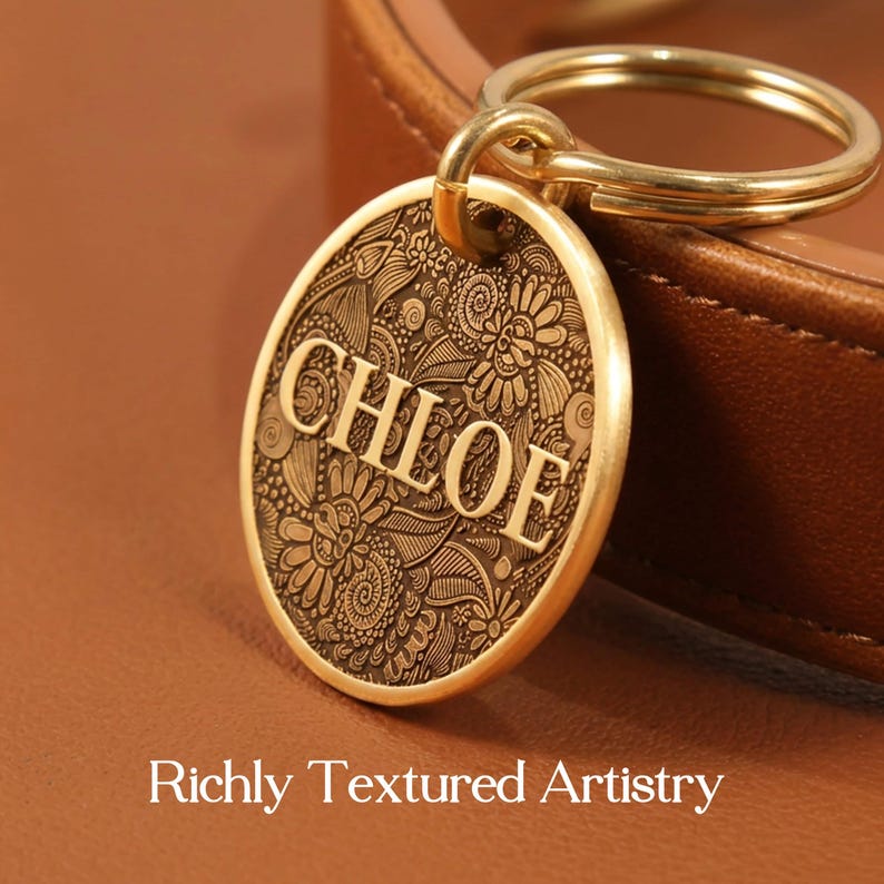 May include: A round gold-toned metal pet tag featuring an intricate engraved floral pattern and the name CHLOE in bold capital letters. The tag is attached to a gold-toned split ring and rests against a smooth brown leather surface.