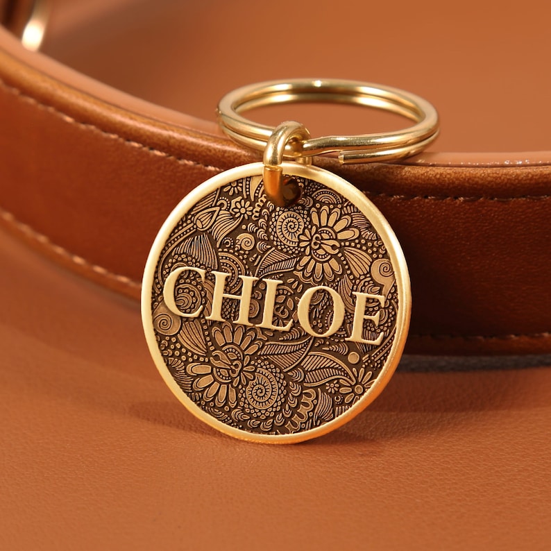 May include: A round, gold-toned metal pet tag featuring an intricate, engraved floral and paisley pattern across its surface. The name CHLOE is embossed in capital letters across the center. The tag is attached to a gold-toned split ring.