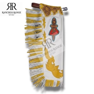 May include: White leather chaps featuring long yellow and white fringe along the side. The upper section has brown floral tooling, while the main body displays a decorative yellow cutout pattern and a central graphic of a dagger with a red heart.