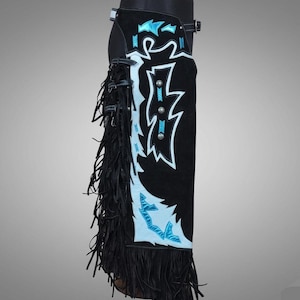 Peut inclure: Chaps en cuir noir avec une finition en daim, présentant un design saisissant en blanc et bleu clair. Les chaps ont une frange sur le côté et des clous décoratifs argentés. Une ceinture et une boucle fixent les chaps à la taille.