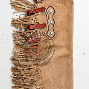 May include: Tan leather chaps with a sunflower design and fringe. Red leather straps and silver-toned embellishments are featured. The logo "TAHIR INDUSTRIES" is visible on the chaps.