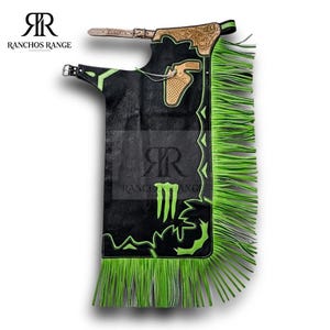 May include: Black leather chaps with neon green fringe and detailing. The chaps display a decorative design and the Monster Energy logo. The top section has a buckle and strap for securing the chaps.