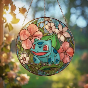 Może przedstawiać: Okrągłe witrażowe okno przedstawiające turkusową postać Bulbasaur otoczoną różowymi i białymi kwiatami oraz zielonymi liśćmi. Okno jest zawieszone na łańcuchu i ma niebieskie tło.
