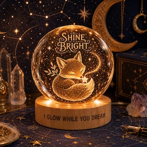 May include: A glowing glass sphere with a sleeping fox design and the words "Shine Bright." The sphere sits on a wooden base that reads "I Glow While You Dream." The fox is holding a star, and the sphere is illuminated with warm light.
