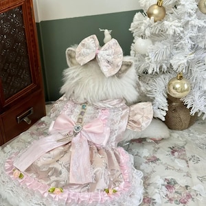 May include: A light pink and white dog dress with a large bow and lace trim. The dress has a satin finish with floral accents and a decorative cross pendant. The dog is wearing a matching bow on its head. The dress is designed for a small dog.