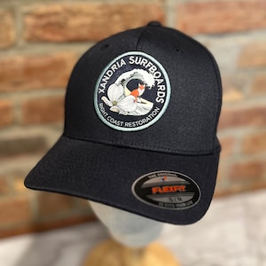 May include: Black Flexfit baseball cap with a circular patch featuring a wave, a fox, and the text "Xandria Surfboards Right Coast Restoration." The cap has a curved brim and a Flexfit tag.
