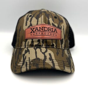 May include: A camouflage trucker hat with a black mesh back. The hat has a copper-colored patch with the text "XANDRIA SURFBOARDS" in black.