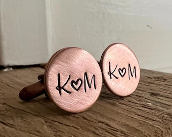 May include: Copper cufflinks with the initials "K♡M" engraved in a handwritten style. The cufflinks are round and have a polished finish. They are displayed on a wooden surface.