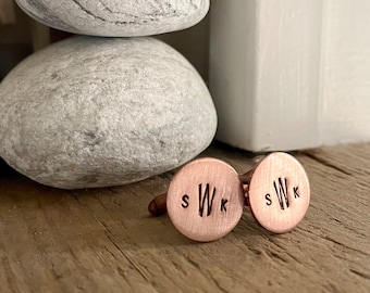 May include: A pair of copper cufflinks with the initials "S W K" engraved on each. The cufflinks are on a wooden surface.
