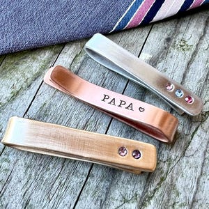 May include: Three metal tie clips in silver, copper, and gold tones. The copper clip is engraved with the word "PAPA" and a heart symbol. The silver clip has three small, colourful gemstones. The gold clip has two gemstones. A striped tie is in the background.