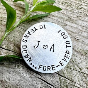 May include: A round, brushed metal keepsake with the text "10 YEARS DOWN...FOREVER TO GO!" and the initials "J ♡ A". Green leaves and a wooden surface provide the background.