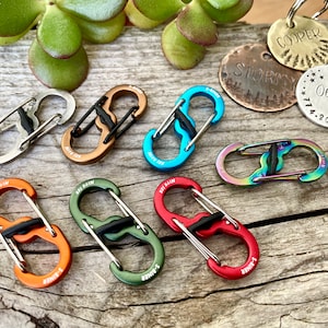 May include: Assortment of eight colorful metal S-biner clips. The clips are in various colors, including silver, bronze, blue, green, orange, and red. The clips are all the same size and shape, and they have a small black plastic piece in the middle.