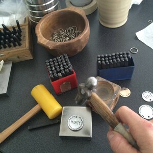 May include: A close-up of a metal stamping set with a hammer and a metal block with the word "MARTY" stamped on it. The set is on a black surface with other tools and supplies.