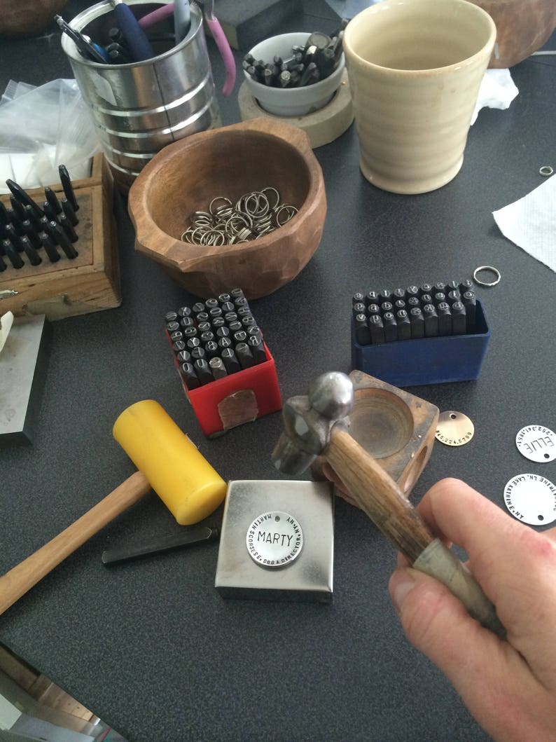 May include: A close-up of a metal stamping set with a hammer and a metal block with the word "MARTY" stamped on it. The set is on a black surface with other tools and supplies.