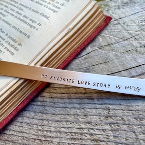 Könnte beinhalten: Ein silbernes Lesezeichen mit dem Text "MY FAVORITE LOVE STORY is ours" eingraviert. Am Ende des Textes ist ein kleines Herz eingraviert.