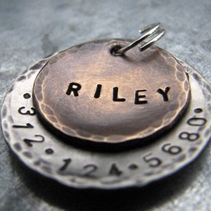 1" Aluminum disc hand stamped with a phone number is behind a 3/4" bronze pet tag stamped with the name "Riley" . Tag hangs from a small split ring.