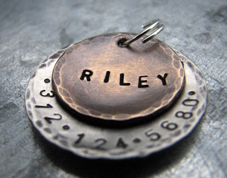 1" Aluminum disc hand stamped with a phone number is behind a 3/4" bronze pet tag stamped with the name "Riley" . Tag hangs from a small split ring.