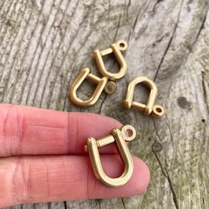 Puede incluir: Primer plano de varios grilletes dorados en forma de U. Un grillete se sostiene en la mano de una persona. Los grilletes tienen un cierre de pasador y un pequeño lazo en la parte superior. El fondo es una superficie de madera desgastada.