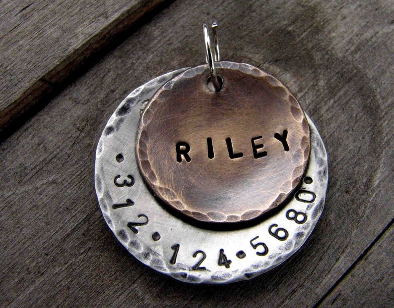 1" Aluminum disc hand stamped with a phone number is behind a 3/4" bronze pet tag stamped with the name "Riley" . Tag hangs from a small split ring.