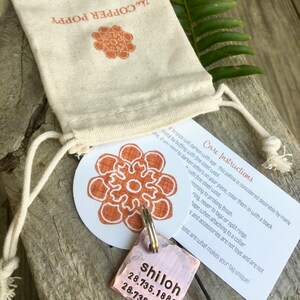 May include: A copper dog tag with the name "shiloh" engraved on it, along with two sets of numbers. The tag is attached to a silver key ring and is sitting on a wooden surface. The tag is accompanied by a small white bag with the words "Copper Poppy" printed on it.