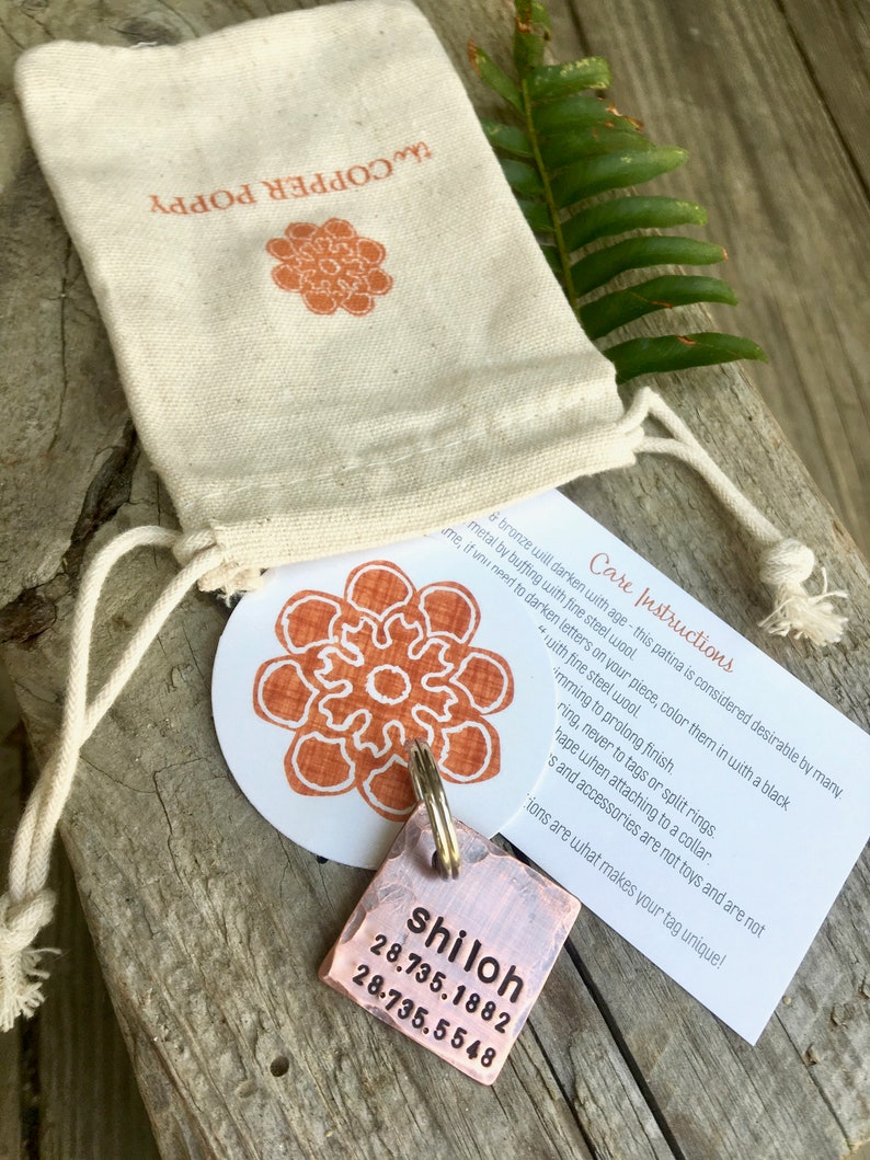 May include: A copper dog tag with the name "shiloh" engraved on it, along with two sets of numbers. The tag is attached to a silver key ring and is sitting on a wooden surface. The tag is accompanied by a small white bag with the words "Copper Poppy" printed on it.