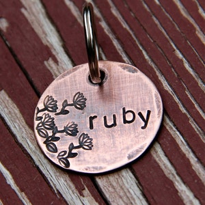 Può includere: Un'etichetta per cani in rame con un motivo floreale e il nome "ruby" inciso su di essa.