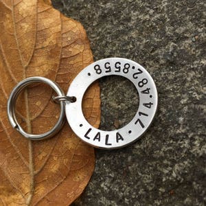May include: A silver dog tag with a round shape and engraved text. The tag has the numbers 8.9.8.2.8.4.7.1.4 and the word "LALA" engraved on it.