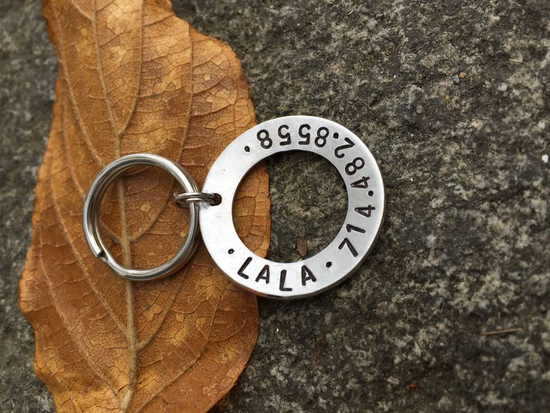 May include: A silver dog tag with a round shape and engraved text. The tag has the numbers 8.9.8.2.8.4.7.1.4 and the word "LALA" engraved on it.