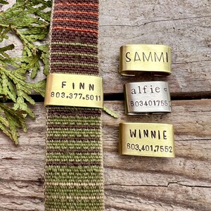 May include: A multi-coloured woven dog collar with brass name tags. The collar features green, brown, and orange tones. The tags are engraved with names and phone numbers: "FINN", "SAMMI", "alfie", and "WINNIE".