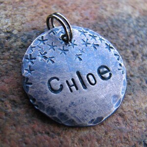 May include: A silver dog tag with a hammered finish and a starburst design around the edge. The name "Chloe" is engraved on the tag.