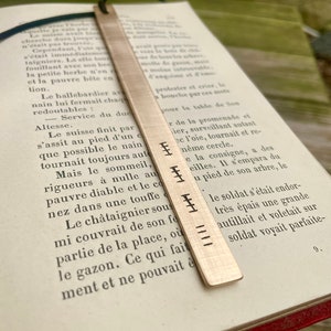 May include: A silver bookmark with a textured surface and engraved lines. The bookmark is placed on a page of a book with text in French.