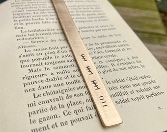 May include: A silver bookmark with a textured surface and engraved lines. The bookmark is placed on a page of a book with text in French.