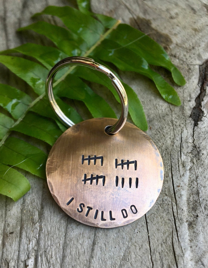 Peut inclure: Un porte-cl&eacute;s rond en laiton avec un anneau de cl&eacute; en argent. Le porte-cl&eacute;s comporte une s&eacute;rie de lignes grav&eacute;es qui repr&eacute;sentent des ann&eacute;es, et les mots "I Still Do" sont grav&eacute;s sous les lignes.