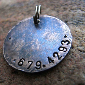 May include: A round, silver-toned pendant with a hammered finish and the numbers 679.4293 engraved on the surface. The pendant has a loop at the top for attaching a chain.