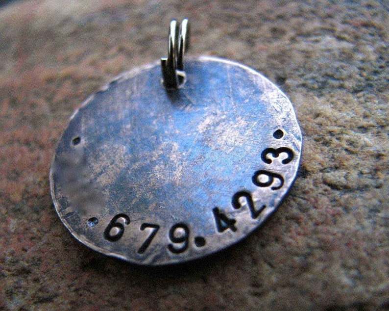 May include: A round, silver-toned pendant with a hammered finish and the numbers 679.4293 engraved on the surface. The pendant has a loop at the top for attaching a chain.