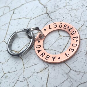 May include: Copper dog tag engraved with the text "DARBY" and "L.966.L.97.036". The tag is attached to a silver key ring.