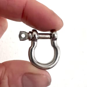 May include: A polished, silver shackle held in a hand. The shackle has a U-shaped body with a pin closure and a small, rectangular opening. It is likely made of stainless steel and is suitable for various applications.