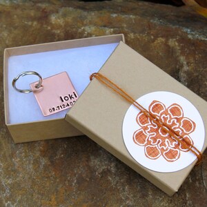 May include: A copper key chain with a personalized tag that reads "Loki 08.7.12.4.02" is displayed in a brown gift box with a white sticker featuring a brown floral design.