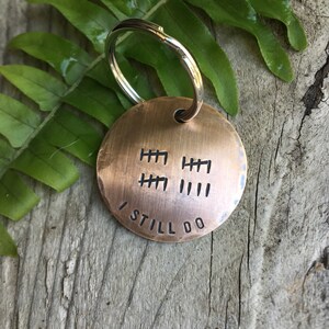 Peut inclure: Un porte-cl&eacute;s rond de couleur cuivre avec un anneau de cl&eacute; argent&eacute;. Le porte-cl&eacute;s comporte une s&eacute;rie de marques de pointage et le texte "I STILL DO" grav&eacute; dessus.
