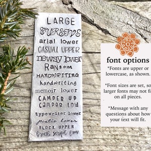 May include: A silver metal tag showcasing font styles such as "LARGE CURSIVE" and "arial lower." A smaller card provides details on font options, sizes, and how to ask about text fitting. The tag is set against a rustic wooden background.