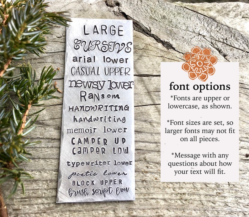May include: A silver metal tag showcasing font styles such as "LARGE CURSIVE" and "arial lower." A smaller card provides details on font options, sizes, and how to ask about text fitting. The tag is set against a rustic wooden background.