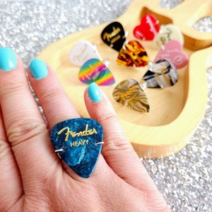 May include: A blue guitar pick ring with the words "Fender Heavy" in gold lettering. The ring is made from a guitar pick and is worn on a finger.