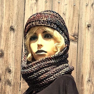 Puede incluir: Un conjunto de gorro y cuello tejido a mano en una mezcla de colores oscuros, incluyendo marrón, negro y rojo. El gorro es de estilo beanie ajustado, y el cuello es una bufanda circular ancha. El conjunto se muestra en un maniquí.