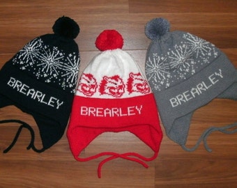 Puede incluir: Tres gorros de invierno de punto con orejeras y pompones. Los gorros son negro, rojo y blanco, y gris. Cada gorro tiene el nombre "Brearley" bordado en la parte delantera.