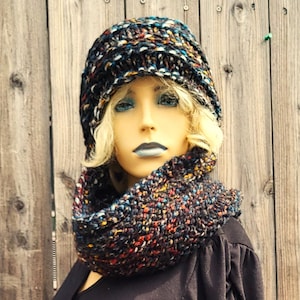 May include: A knitted hat and cowl set in a dark yarn with colourful flecks. The beanie hat and circular scarf are displayed on a mannequin. The yarn includes flecks of blue, red, yellow, and white, creating a textured effect.