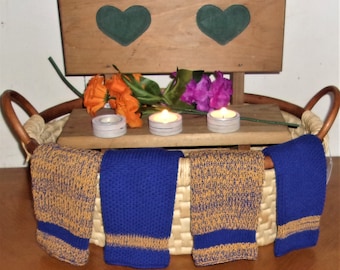 May include: A wicker basket with four knitted dishcloths. Two are blue with a gold stripe and two are gold with a blue stripe. There are three white candles and a bouquet of orange and purple flowers on a wooden shelf above the basket.