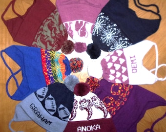 Puede incluir: Una colección de ocho gorros de punto con orejeras dispuestos en círculo, con varios colores y patrones que incluyen balones de fútbol, corazones y motivos animales. Algunos gorros están personalizados con los nombres Graham, Demi y Anoka.