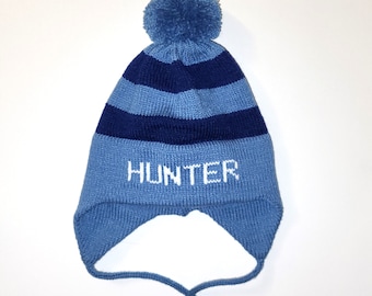 Puede incluir: Un gorro de punto con orejeras y un pompón en la parte superior, con rayas horizontales en azul claro y azul marino. La palabra HUNTER está tejida en letras blancas en la parte frontal. Dos finas cintas cuelgan de las orejeras.